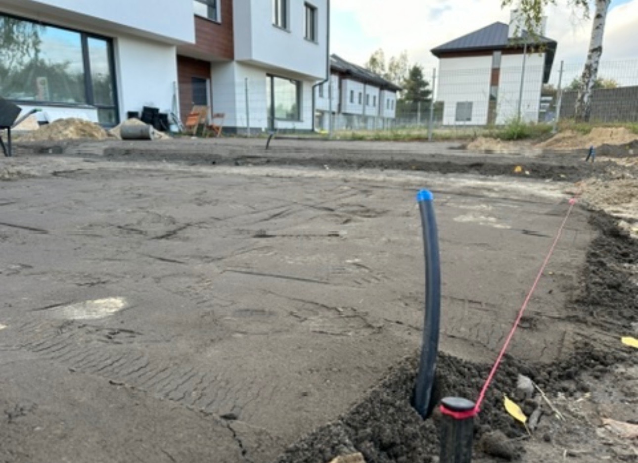 Przygotowany teren pod trawnik z widocznymi rurami nawadniającymi i wytyczonym obrysem. W tle budynki mieszkalne. Etap prac ziemnych przed założeniem trawnika.