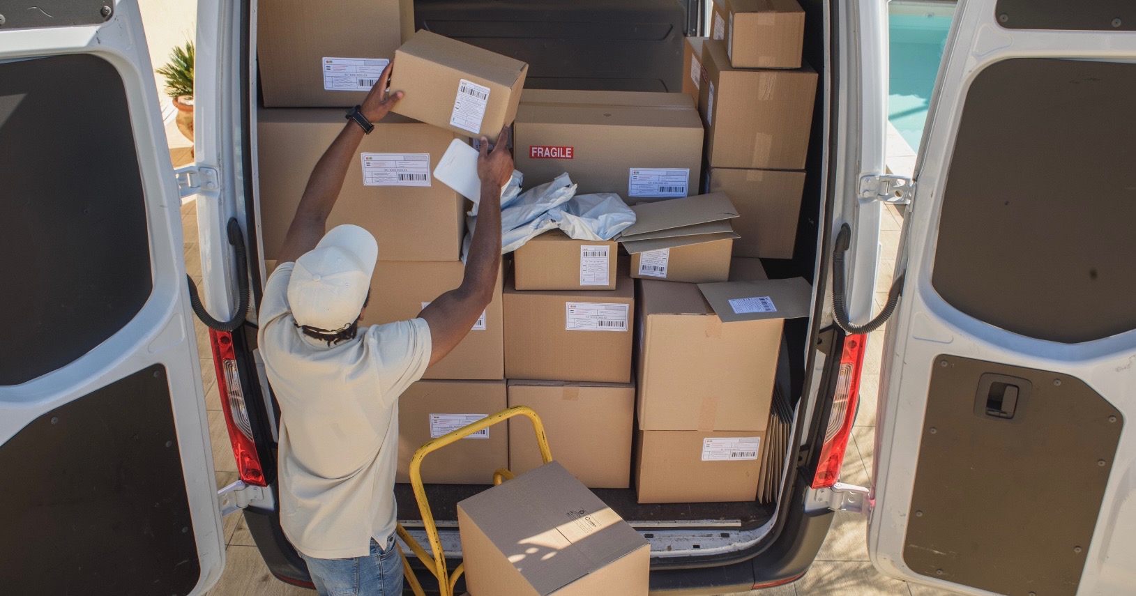 Osoba w białej czapce i koszulce wkłada kartonowe pudła do białego busa. Widoczny żółty wózek transportowy i pudełka z napisem 'Fragile'.