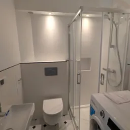 Nowoczesna łazienka w bieli z kabiną prysznicową, umywalką i toaletą. Na podłodze białe płytki z czarnymi akcentami. Pralka częściowo widoczna po prawej stronie.