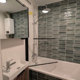 Leonidas & I-BUD - Nowoczesna łazienka z wanną obłożoną szarymi, podłużnymi płytkami, białą umywalką z szafką z drewnianymi lamelami i srebrną armaturą. Widoczny bojler.