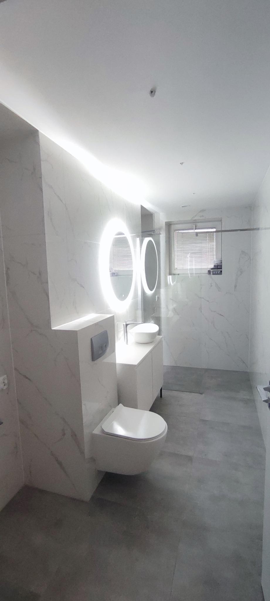 Nowoczesna łazienka w bieli i szarości. Umywalka z szafką, podwieszana toaleta i okrągłe lustro z podświetleniem LED. Marmurowe płytki na ścianach i szara podłoga.