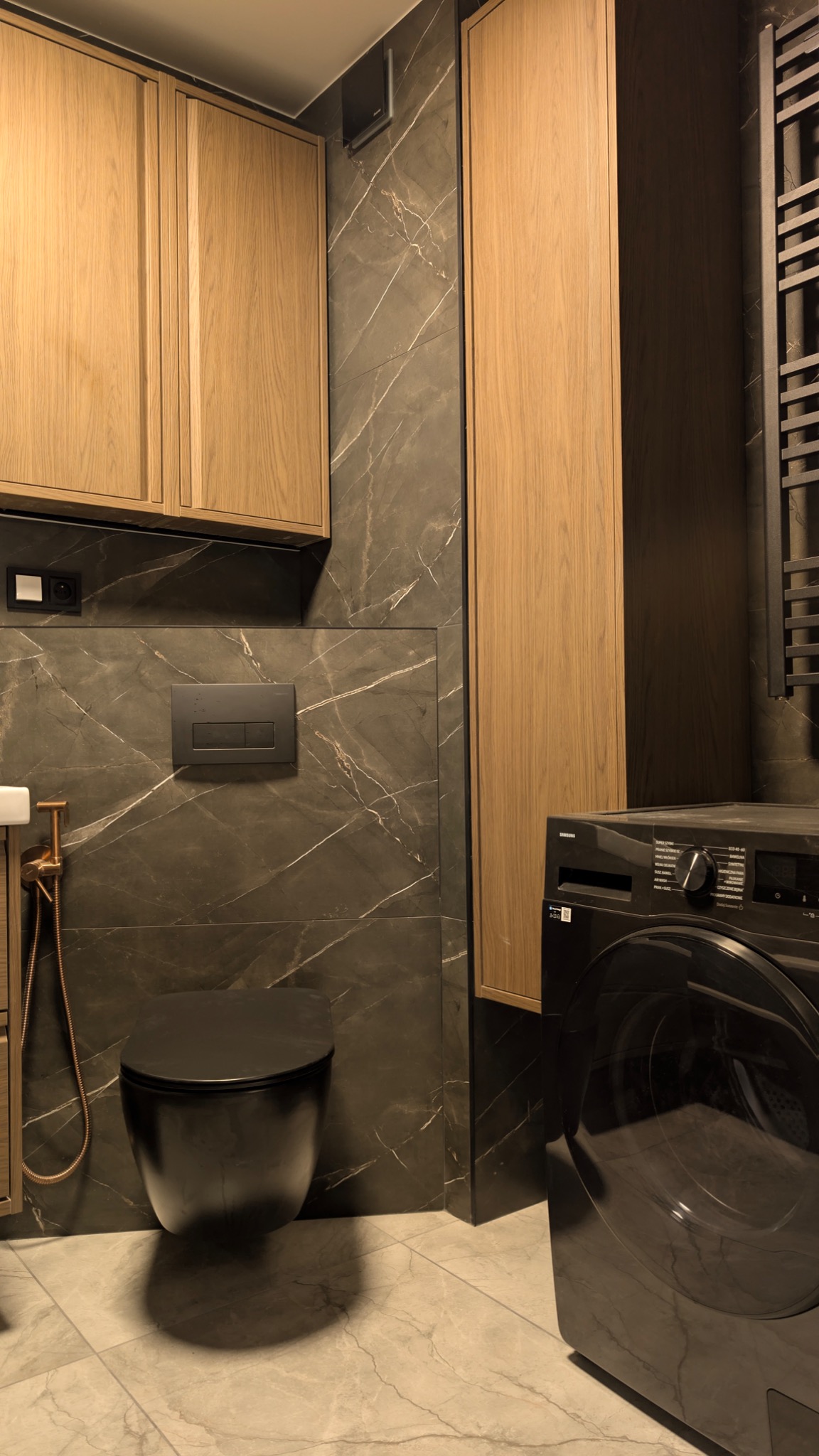 Nowoczesna łazienka z marmurowymi płytkami, wiszącą toaletą i pralką Samsung. Uwagę przykuwają drewniane szafki i złota armatura.