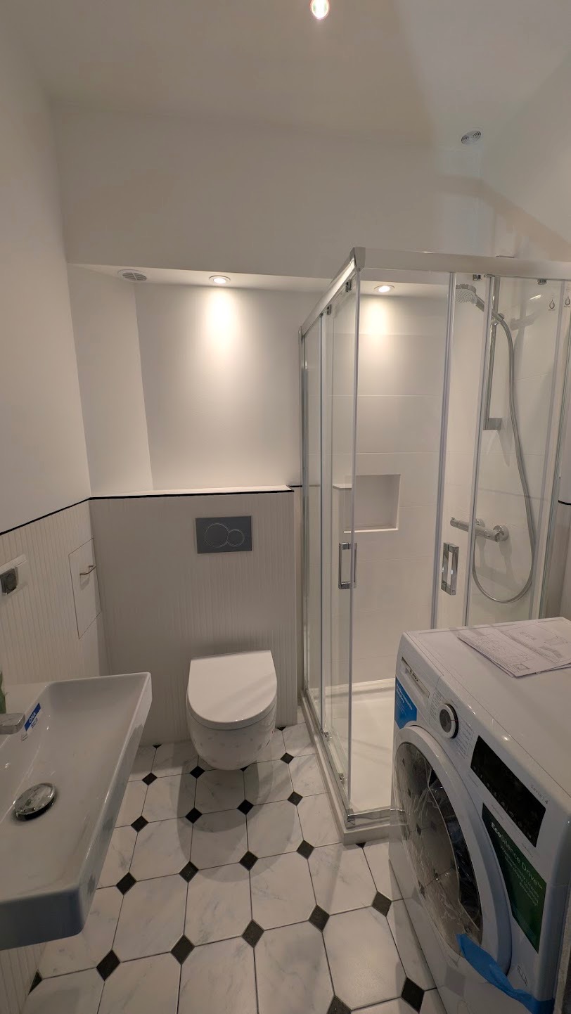 Nowoczesna łazienka w bieli z kabiną prysznicową, umywalką i toaletą. Na podłodze białe płytki z czarnymi akcentami. Pralka częściowo widoczna po prawej stronie.