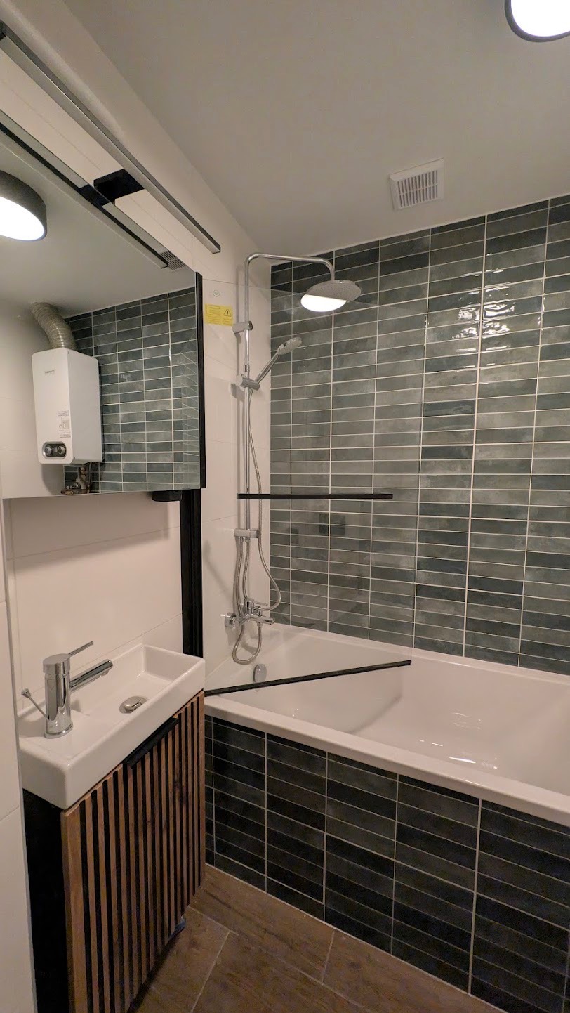 Nowoczesna łazienka z wanną obłożoną szarymi, podłużnymi płytkami, białą umywalką z szafką z drewnianymi lamelami i srebrną armaturą. Widoczny bojler.