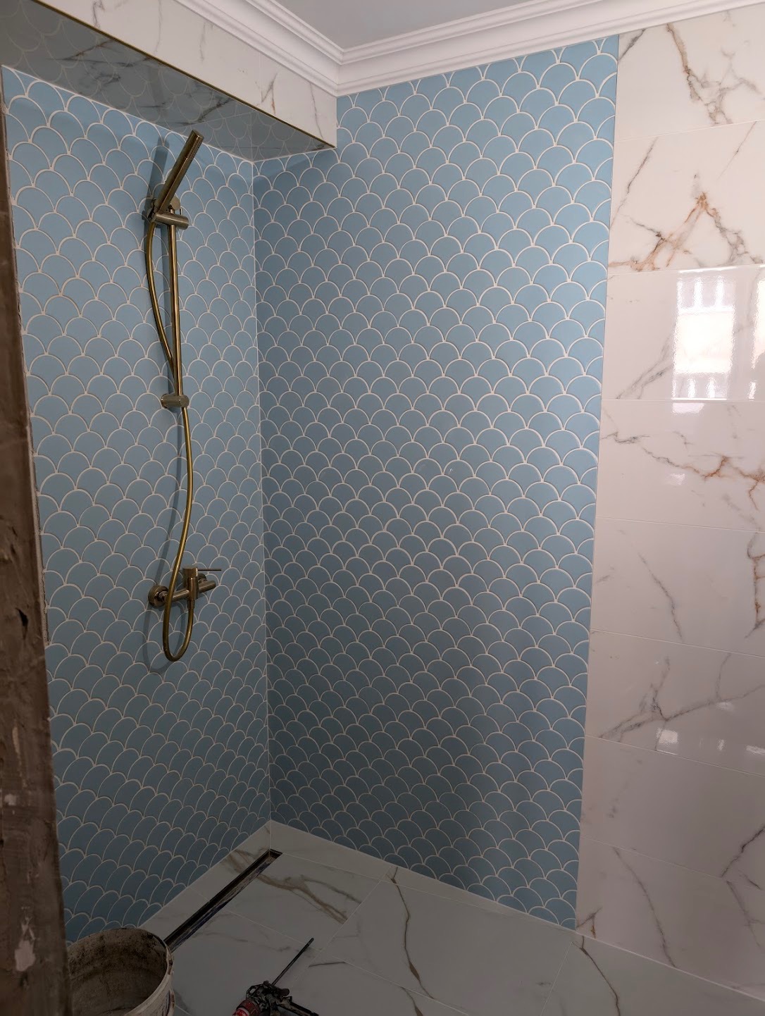 Wykończona łazienka z błękitnymi płytkami w kształcie rybiej łuski i białymi płytkami imitującymi marmur. Widoczny złoty zestaw prysznicowy i odpływ liniowy w podłodze.