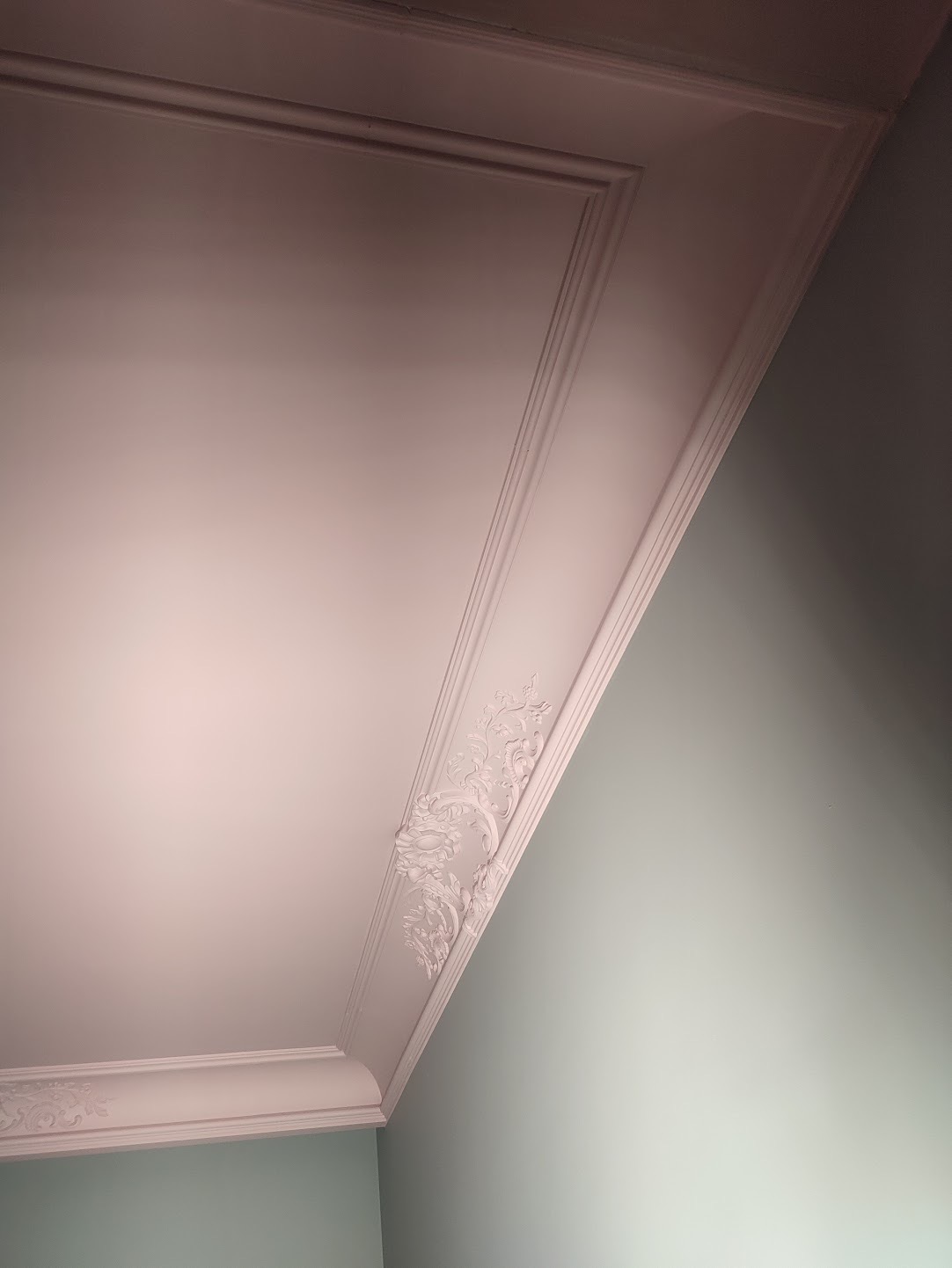 Biały sufit z ozdobną, gipsową sztukaterią w rogu pokoju, widok pod kątem. Ściana w odcieniu stonowanej zieleni. Detal wykończenia wnętrza.