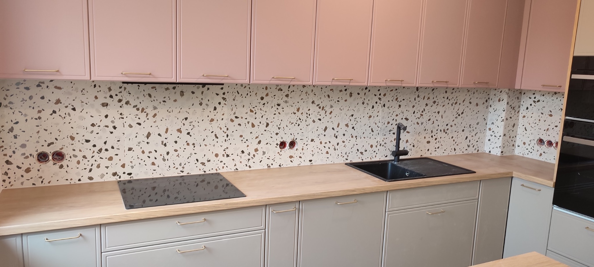 Nowoczesna kuchnia z różowymi szafkami górnymi i mozaikową ścianą nad blatem. Widoczne płyta indukcyjna i zlew. Neutralna kolorystyka dolnych szafek.