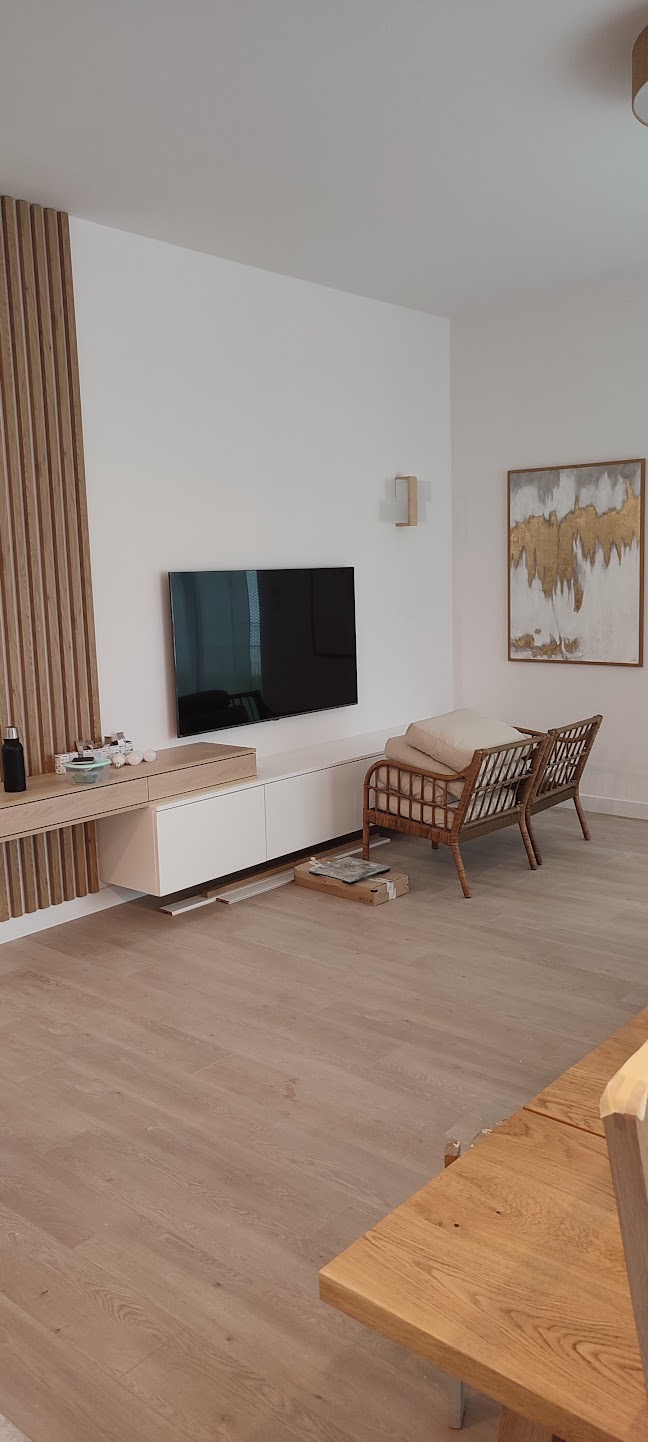 Nowoczesny salon z drewnianą ścianą, TV, białymi szafkami RTV i rattanowymi fotelami. Jasna podłoga i obraz w złotej ramie dopełniają wystrój.