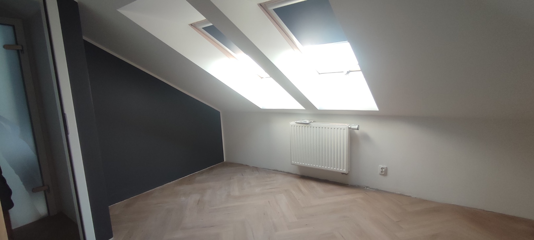 Pokój na poddaszu po remoncie: ciemna ściana, jasna podłoga w jodełkę, okna dachowe z białymi ramami, biały grzejnik. Widoczny kontakt elektryczny.
