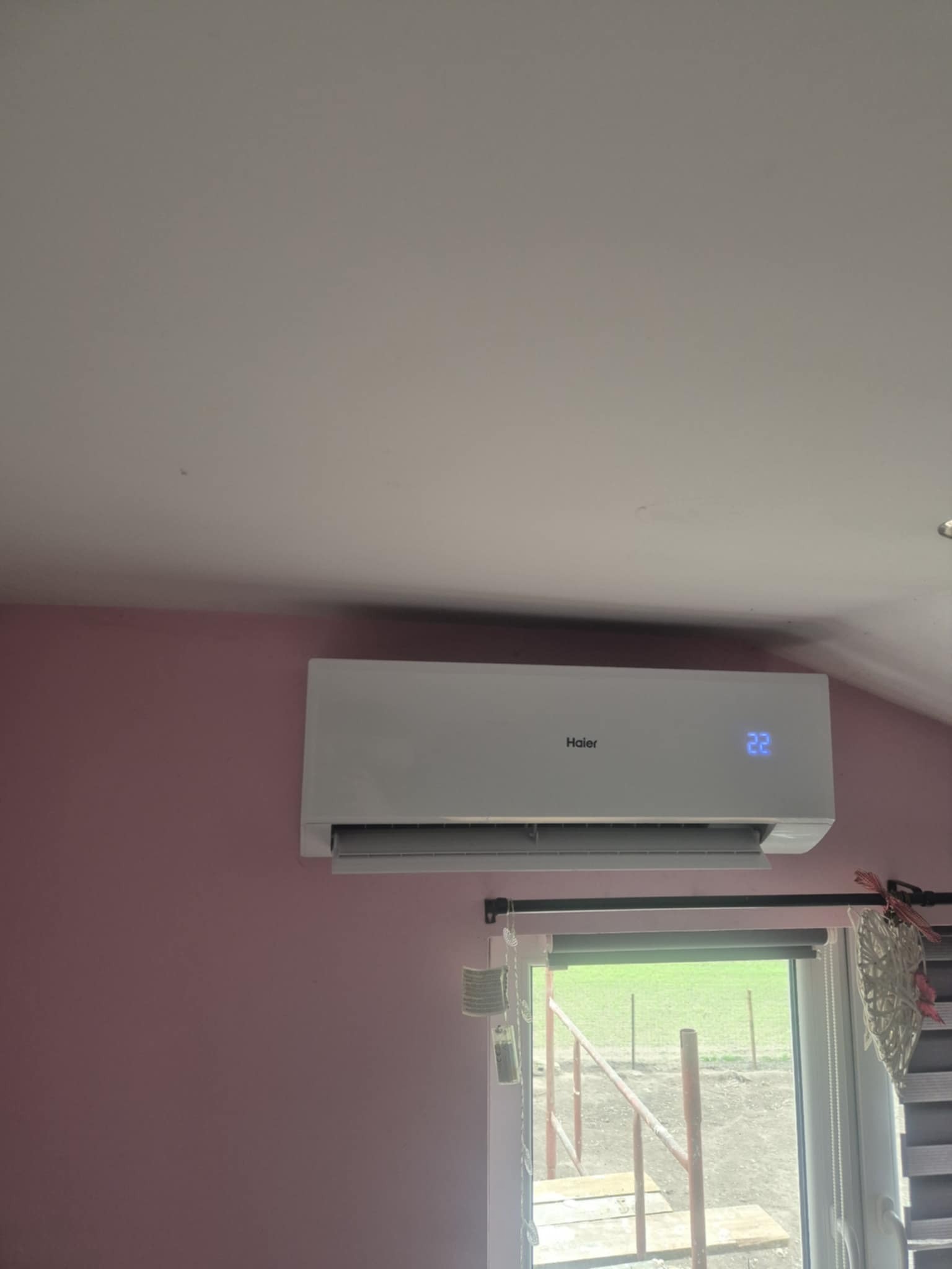 Biała klimatyzacja Haier na ścianie w pokoju, ustawiona na 22 stopnie, widok przez okno na zewnątrz.