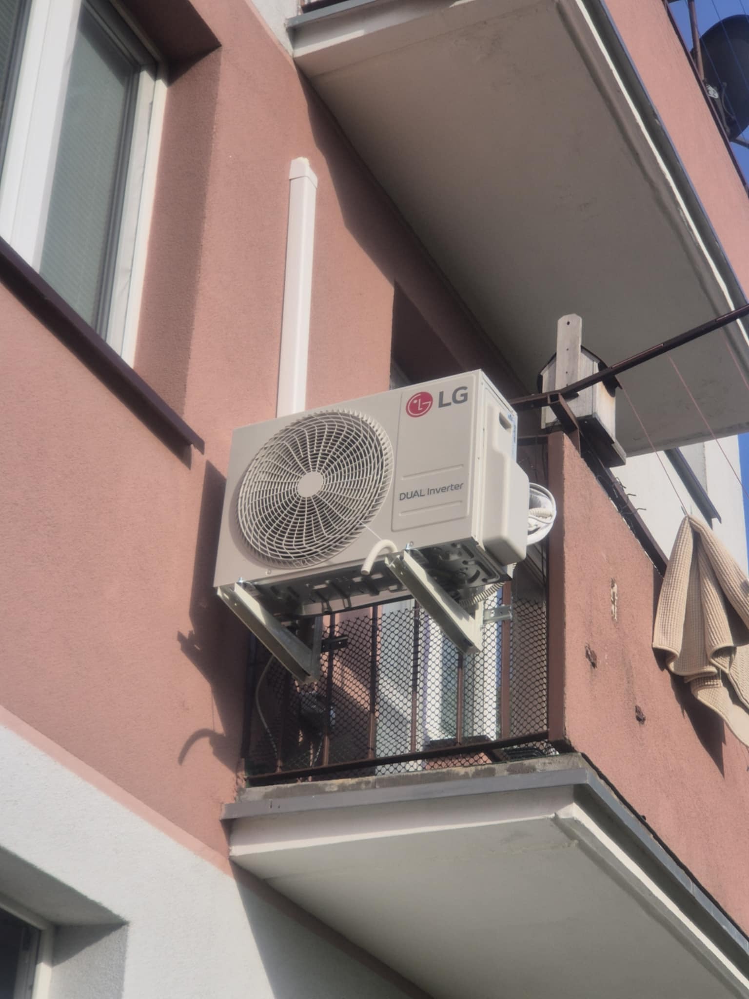 Zewnętrzna jednostka klimatyzacji LG Dual Inverter zamontowana na balkonie bloku mieszkalnego. Widoczny fragment elewacji budynku i barierka balkonu.