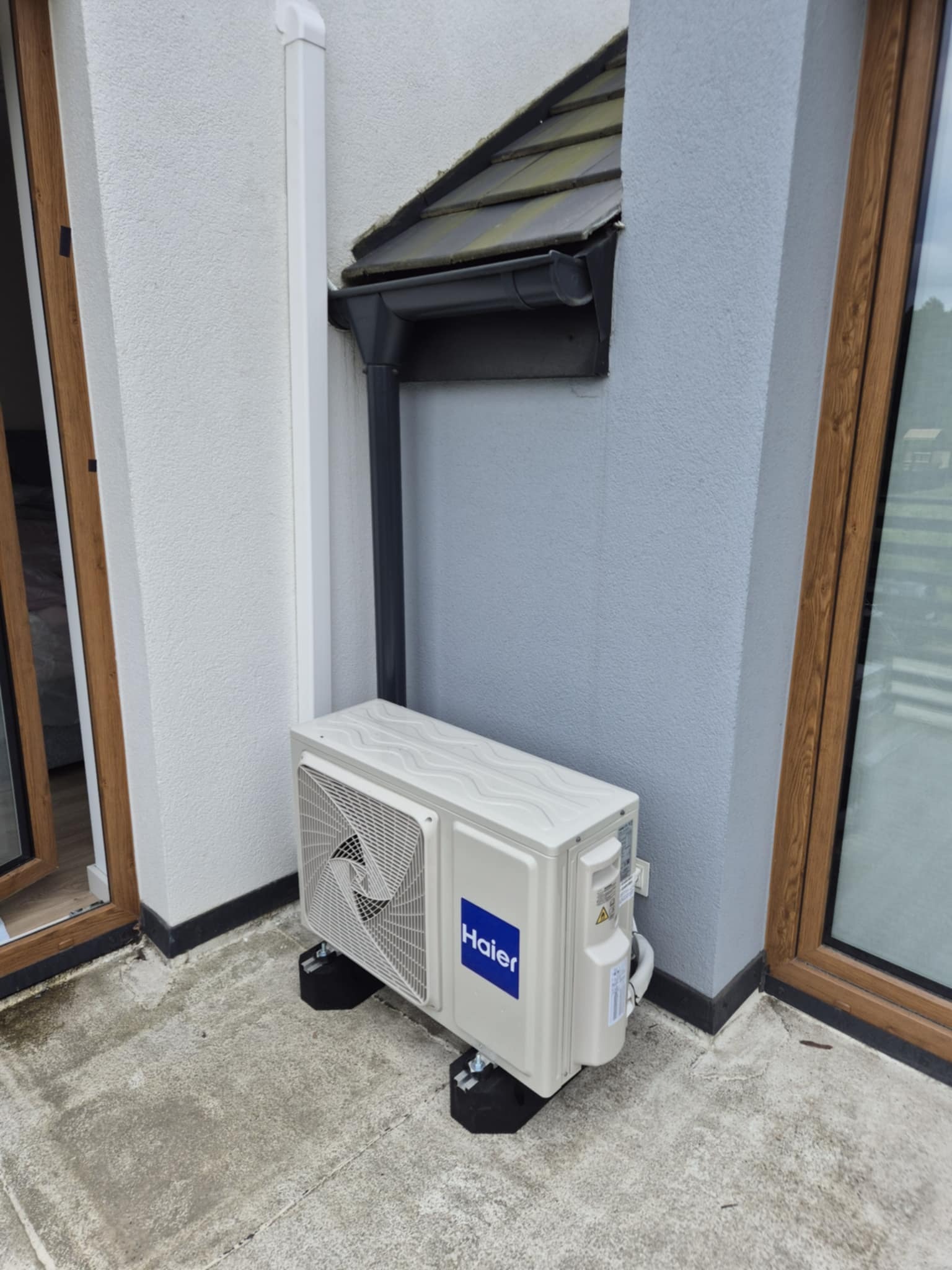 Biały klimatyzator Haier zamontowany na zewnątrz budynku, pod rynną. Widoczne czarne podkładki pod urządzeniem i fragmenty okien z drewnianą ramą.