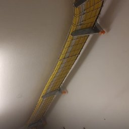 Mateusz Darski ENERGY GUARDS - Koryto kablowe z żółtymi i białymi kablami, zamontowane pod sufitem, widok z dołu. Metalowe wsporniki mocują koryto do sufitu. Instalacja wewnątrz budynku.