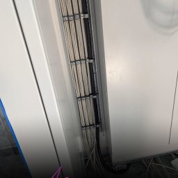 Mateusz Darski ENERGY GUARDS - Równolegle ułożone kable elektryczne w metalowych uchwytach, prowadzone wzdłuż białej ściany. Widoczne mocowania i fragmenty instalacji.