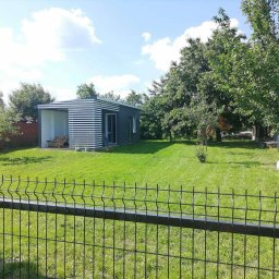 Perfect-Color - Nowoczesny domek narzędziowy z białymi poziomymi panelami, zielony trawnik i metalowe ogrodzenie panelowe. Osoba pracuje w ogrodzie za drzewem.