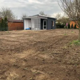 Przygotowany teren pod założenie ogrodu z domkiem narzędziowym w tle. Wyrównana ziemia, widoczne ślady po bronowaniu. Pochmurne niebo.