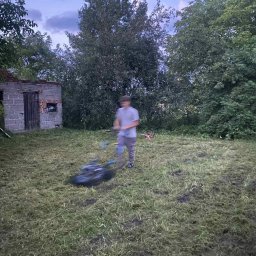 Perfect-Color - Mężczyzna kosi trawę na zewnątrz, obok starego, zniszczonego budynku. Widoczne ślady po koszeniu trawy i resztki skoszonej trawy na trawniku.