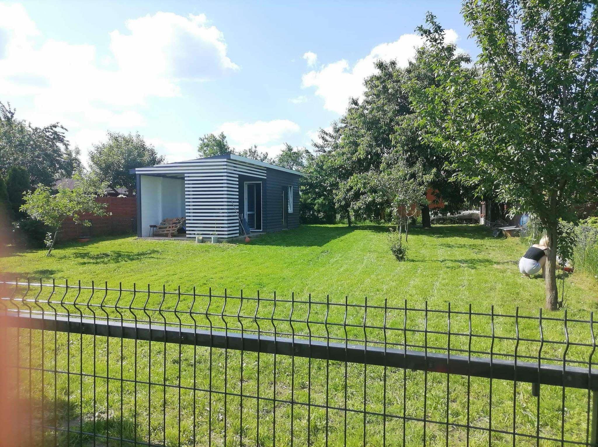 Nowoczesny domek narzędziowy z białymi poziomymi panelami, zielony trawnik i metalowe ogrodzenie panelowe. Osoba pracuje w ogrodzie za drzewem.