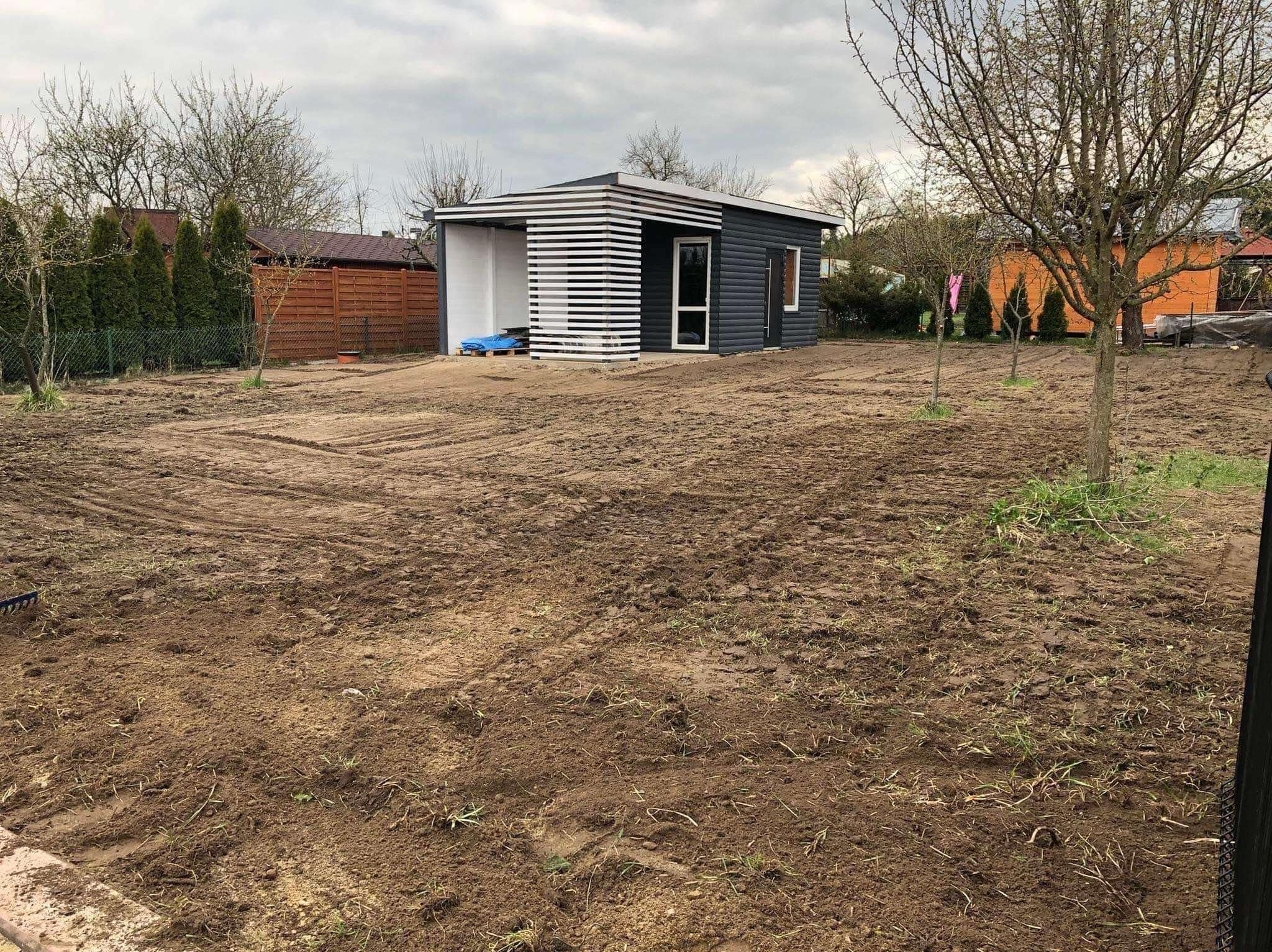 Przygotowany teren pod założenie ogrodu z domkiem narzędziowym w tle. Wyrównana ziemia, widoczne ślady po bronowaniu. Pochmurne niebo.