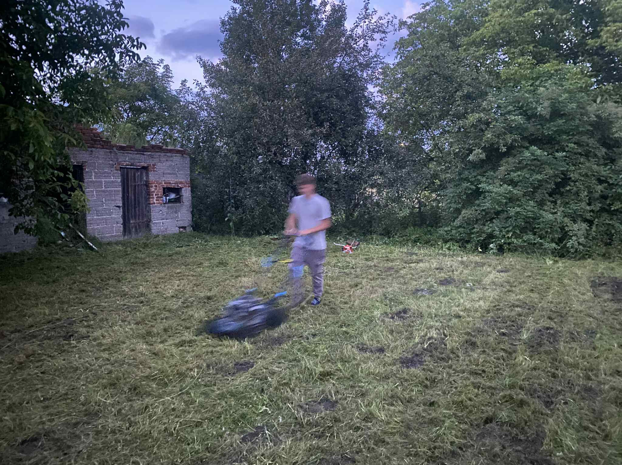 Mężczyzna kosi trawę na zewnątrz, obok starego, zniszczonego budynku. Widoczne ślady po koszeniu trawy i resztki skoszonej trawy na trawniku.
