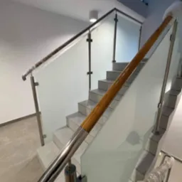 Szklana balustrada z drewnianym i metalowym wykończeniem na schodach w jasnym wnętrzu. Elementy balustrady zabezpieczone folią ochronną.