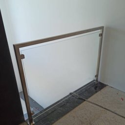 SKs - Szklana balustrada z metalową ramą zamontowana na granicy dwóch poziomów posadzki. Widoczne mocowania i elementy konstrukcyjne. Minimalistyczny design.