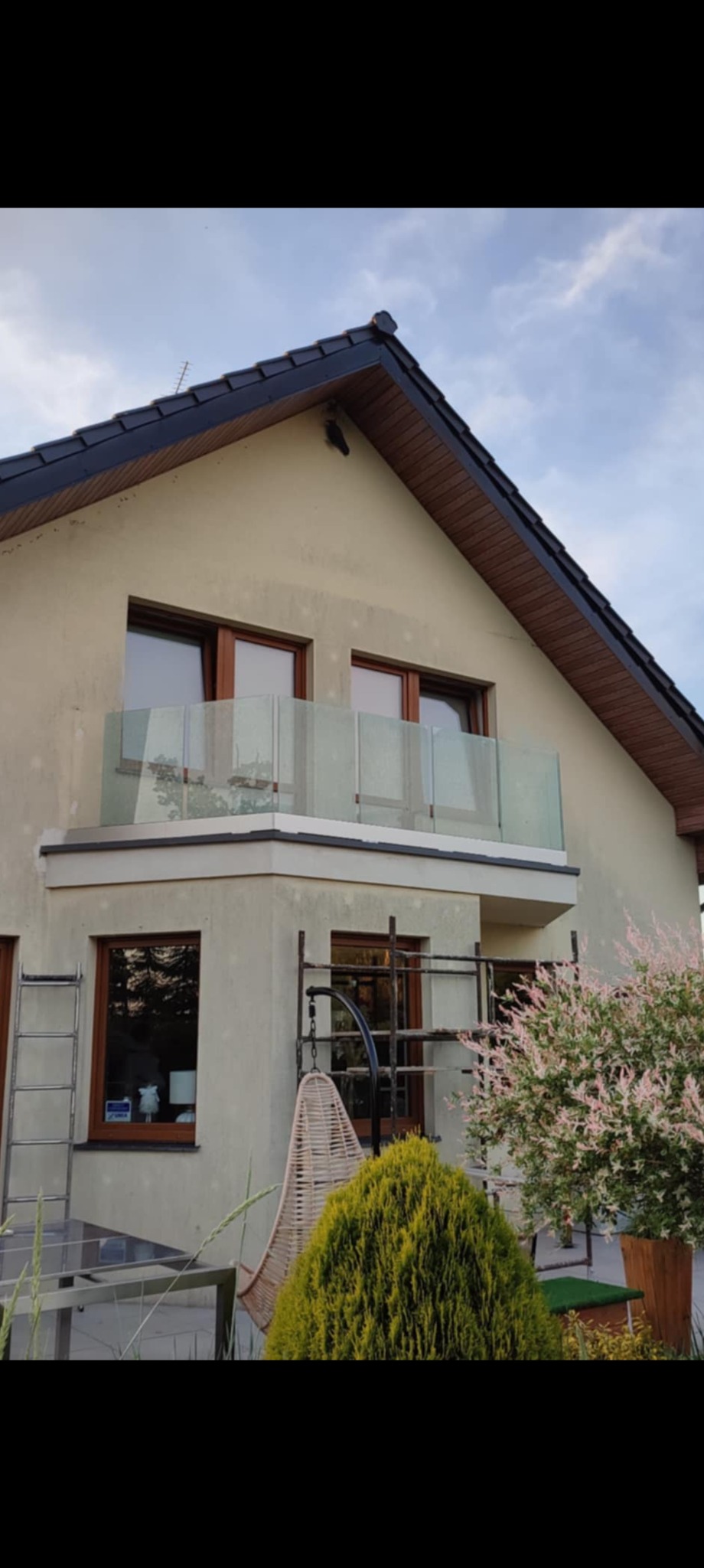 Dom z balkonem zabezpieczonym szklaną balustradą, widoczne rusztowanie i drabina, sugerujące prace wykończeniowe.