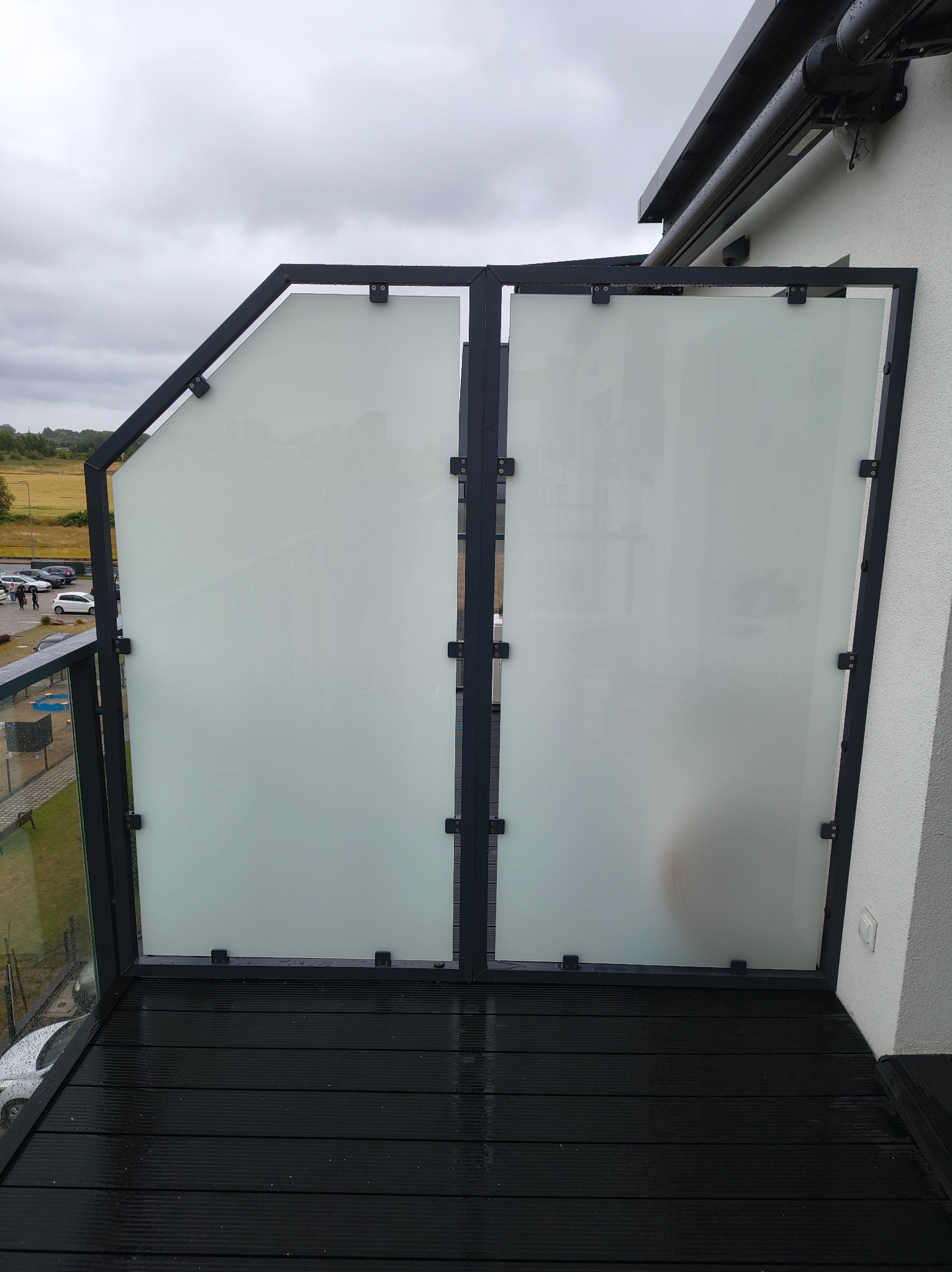 Szklana balustrada balkonowa z czarną, metalową ramą i matowym szkłem, widok z góry na mokrą, drewnianą podłogę balkonu. Minimalistyczny design.