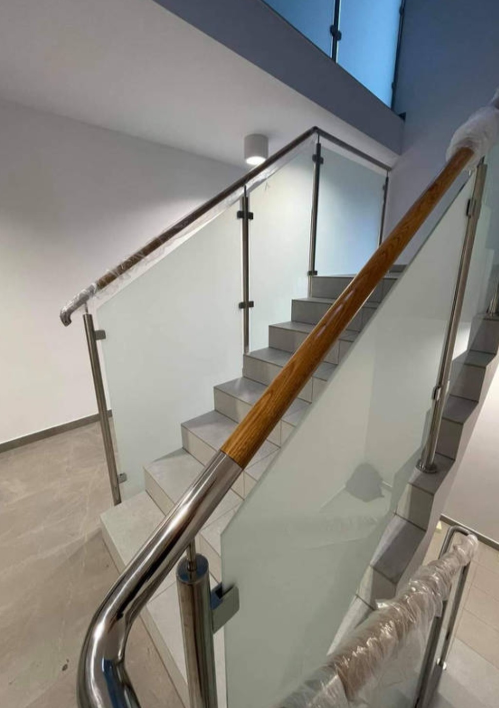 Szklana balustrada z drewnianym i metalowym wykończeniem na schodach w jasnym wnętrzu. Elementy balustrady zabezpieczone folią ochronną.