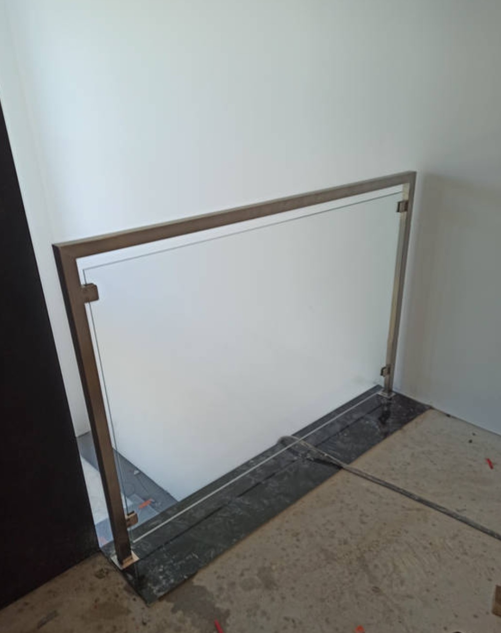 Szklana balustrada z metalową ramą zamontowana na granicy dwóch poziomów posadzki. Widoczne mocowania i elementy konstrukcyjne. Minimalistyczny design.