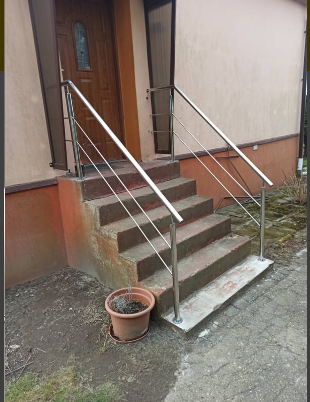 Zewnętrzne schody z brązowymi stopniami i nowoczesną, metalową, srebrną balustradą. Obok schodów stoi doniczka z rośliną. Widok na brązowe drzwi wejściowe.