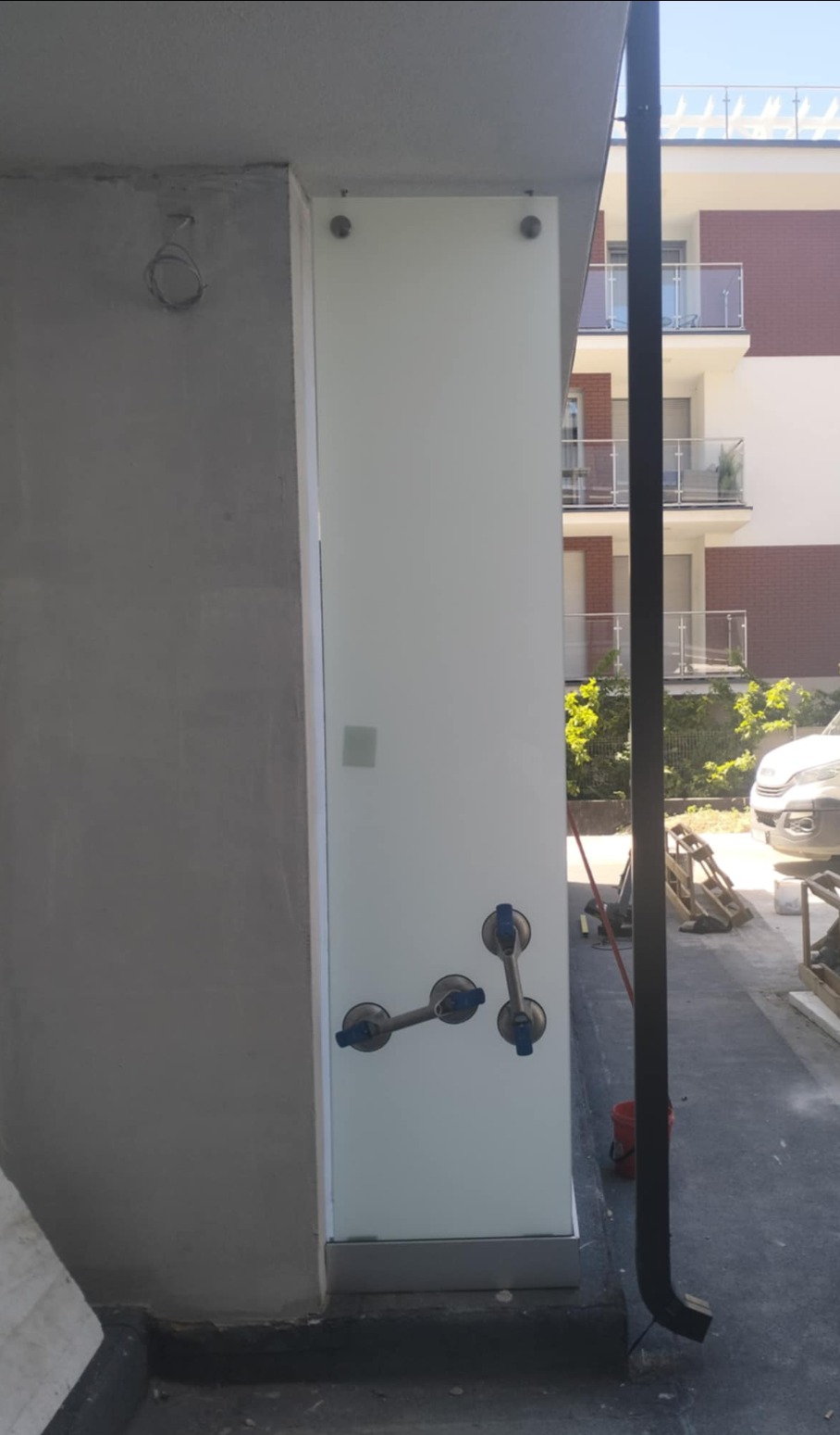 Szklana tafla mocowana przyssawkami do budynku. Widoczne otwory na śruby. W tle budynek wielorodzinny z balkonami.