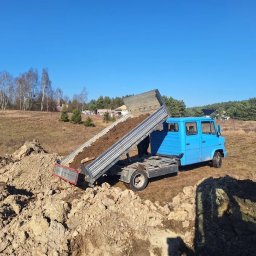 ARBA KAROLINA SŁABIG - Niebieska wywrotka z podniesioną skrzynią ładunkową wysypuje ziemię na placu budowy, w tle drzewa i błękitne niebo.