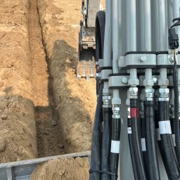 ARBA KAROLINA SŁABIG - Koparka w trakcie prac ziemnych. Widoczny wykop w piaszczystej glebie i operator w oddali. Detal hydrauliki maszyny na pierwszym planie.