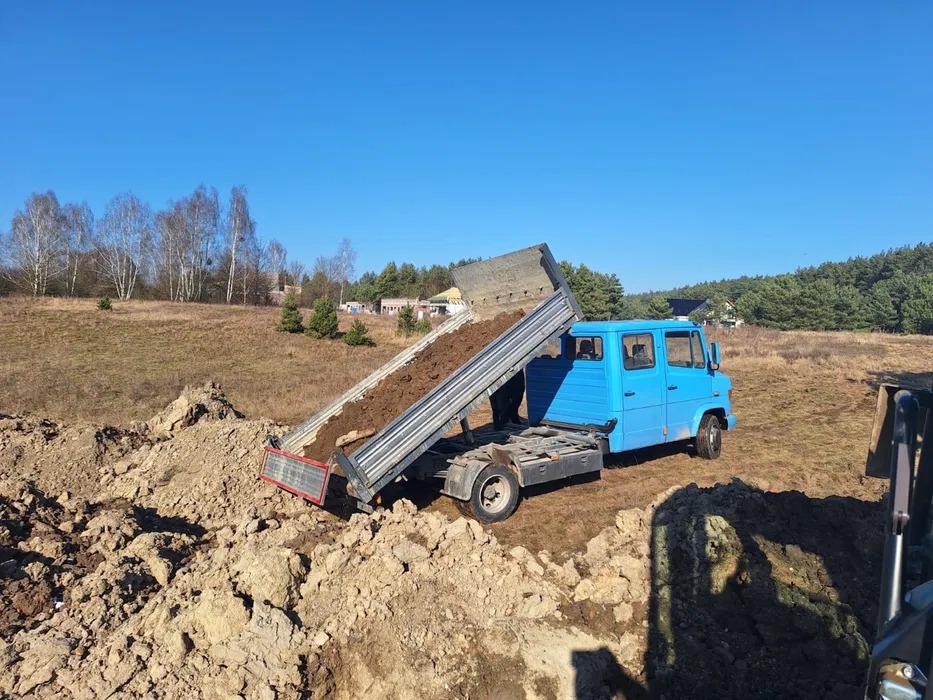 Niebieska wywrotka z podniesioną skrzynią ładunkową wysypuje ziemię na placu budowy, w tle drzewa i błękitne niebo.