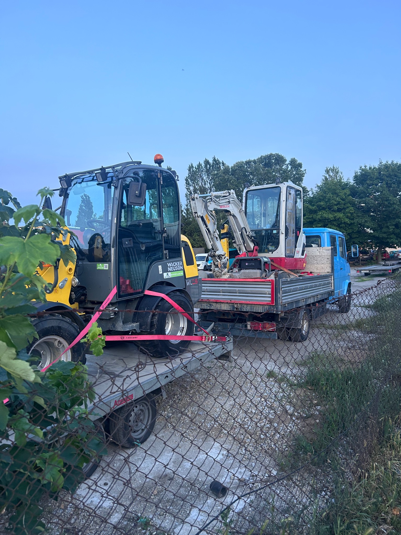 Transport małej koparki i ładowarki na przyczepie i samochodzie ciężarowym. Widok przez siatkę ogrodzeniową, zieleń w tle. Sprzęt budowlany.