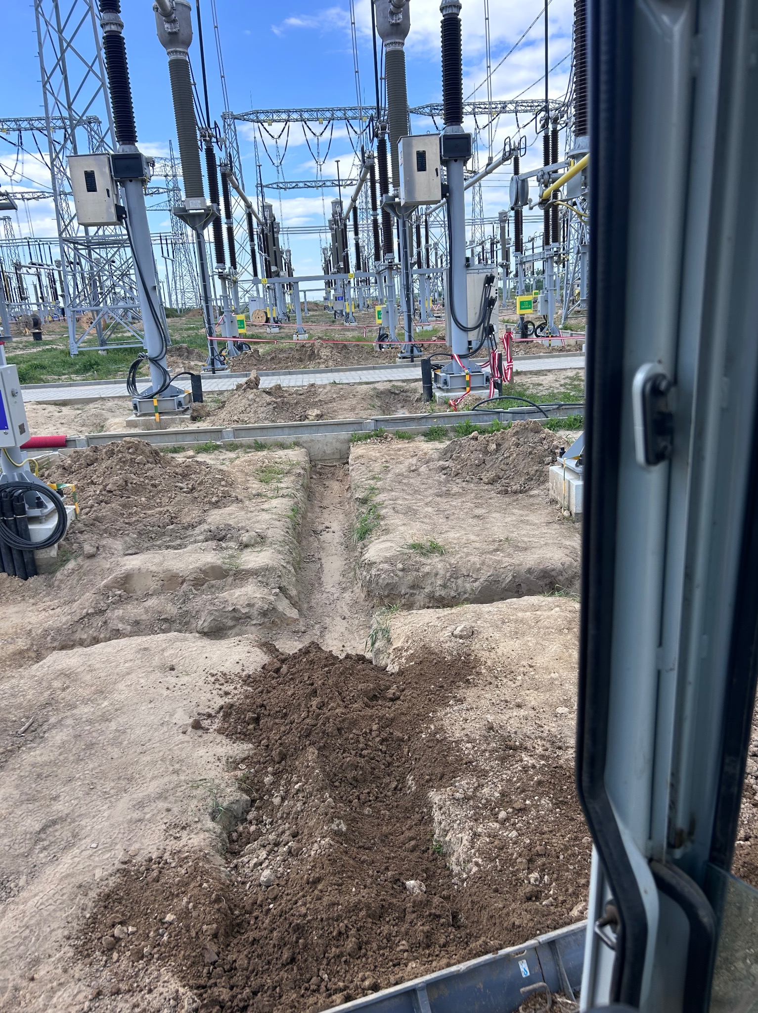 Wykopany wąski rów w ziemi na terenie stacji transformatorowej, widok z kabiny maszyny. Rów przygotowany pod instalacje, z hałdami ziemi po bokach.