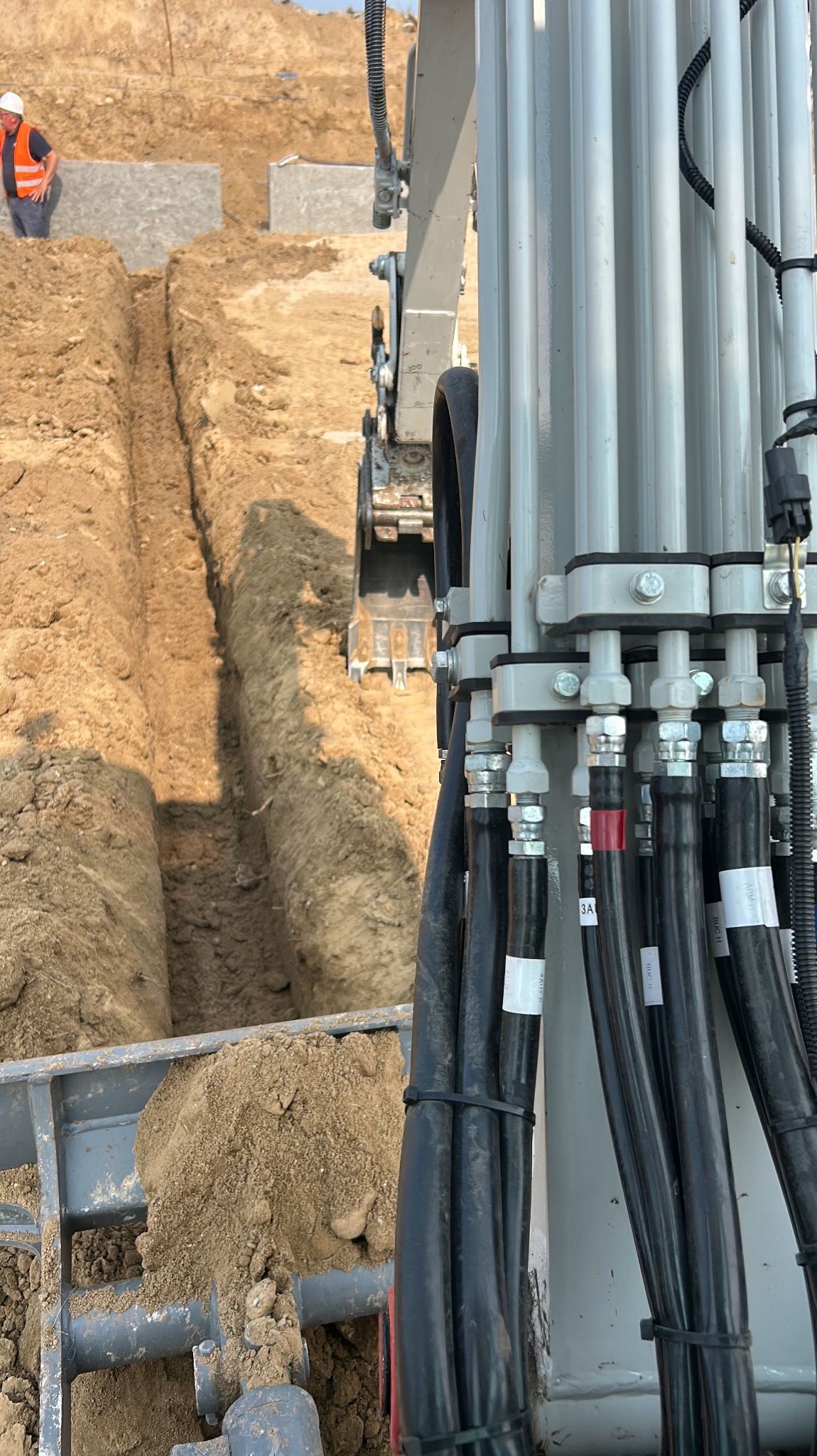 Koparka w trakcie prac ziemnych. Widoczny wykop w piaszczystej glebie i operator w oddali. Detal hydrauliki maszyny na pierwszym planie.