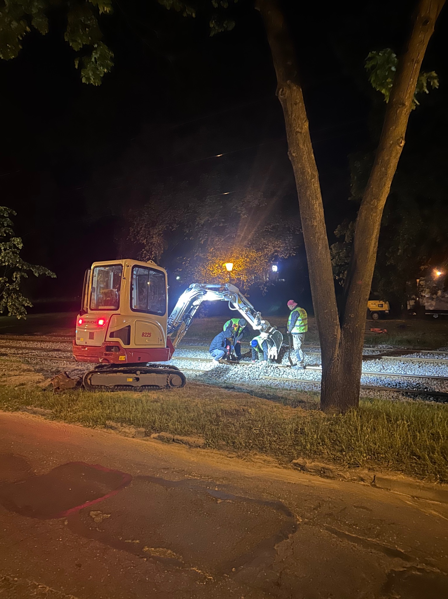 Prace ziemne nocą: mini-koparka Takeuchi B225 oświetla grupę robotników w kamizelkach odblaskowych, pracujących na nasypie kolejowym. W tle widoczne drzewa i oświetlenie uliczne.