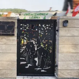 Czarna, metalowa brama wjazdowa z wycinanym motywem wilków na tle drzew, zamontowana na jasnych, kamiennych słupkach. Widoczna kostka brukowa i budynek w tle.