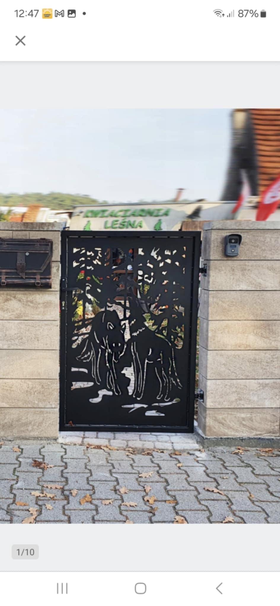 Czarna, metalowa brama wjazdowa z wycinanym motywem wilków na tle drzew, zamontowana na jasnych, kamiennych słupkach. Widoczna kostka brukowa i budynek w tle.