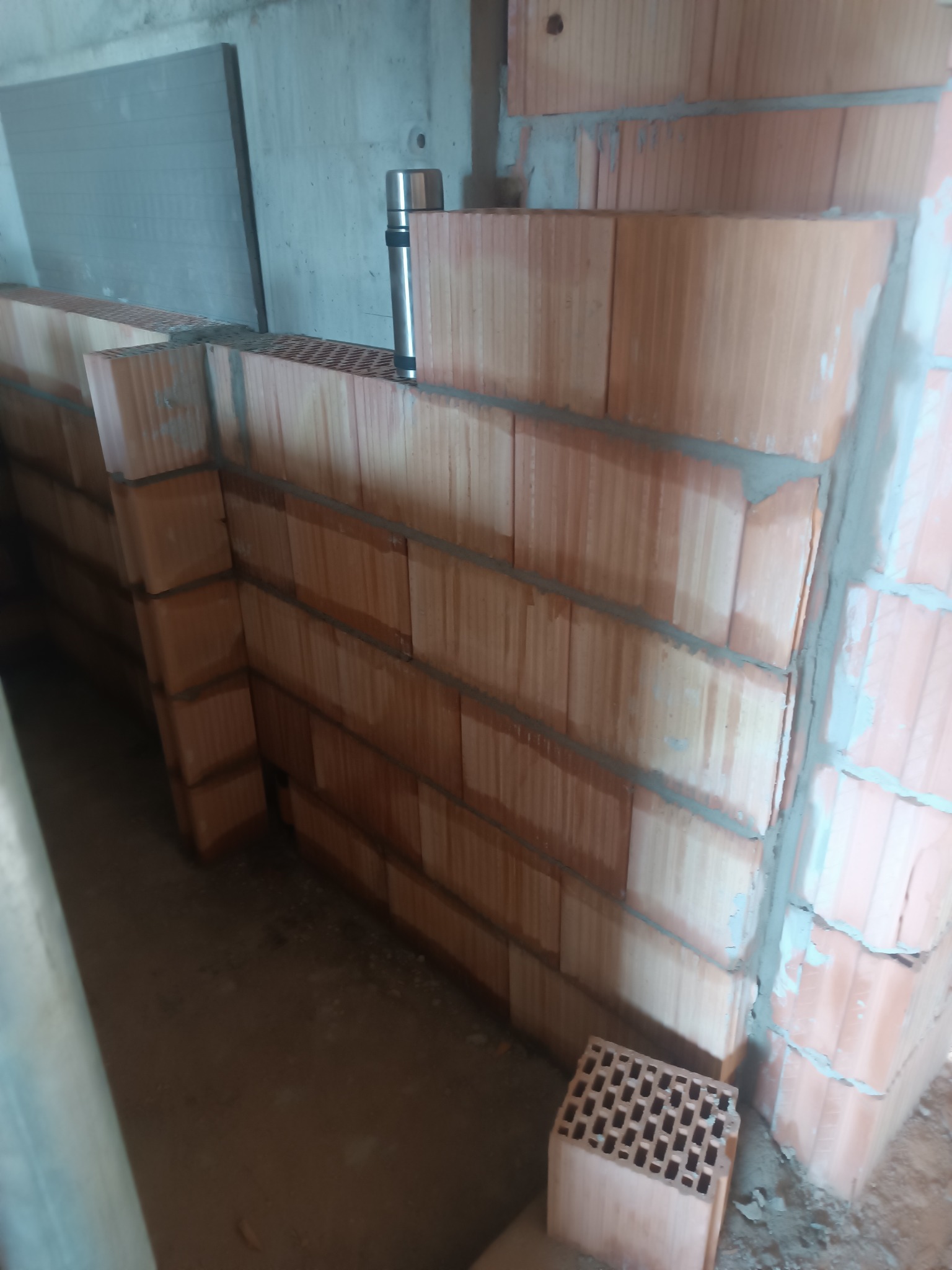 Wznoszenie ściany z pustaków ceramicznych, widoczne spoiny z zaprawy. Na murze stoi termos, a obok leży pojedynczy pustak. Surowe wnętrze w trakcie budowy.