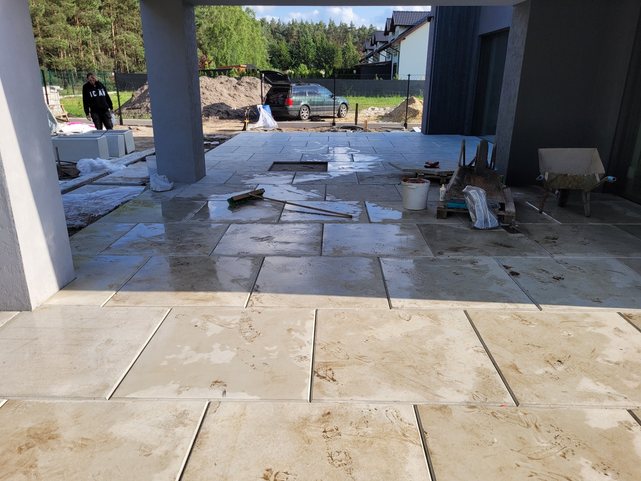 Jasne, kwadratowe płyty tarasowe w trakcie montażu, częściowo mokre, z widocznymi narzędziami i pracownikiem w tle, na tle zieleni i domu.