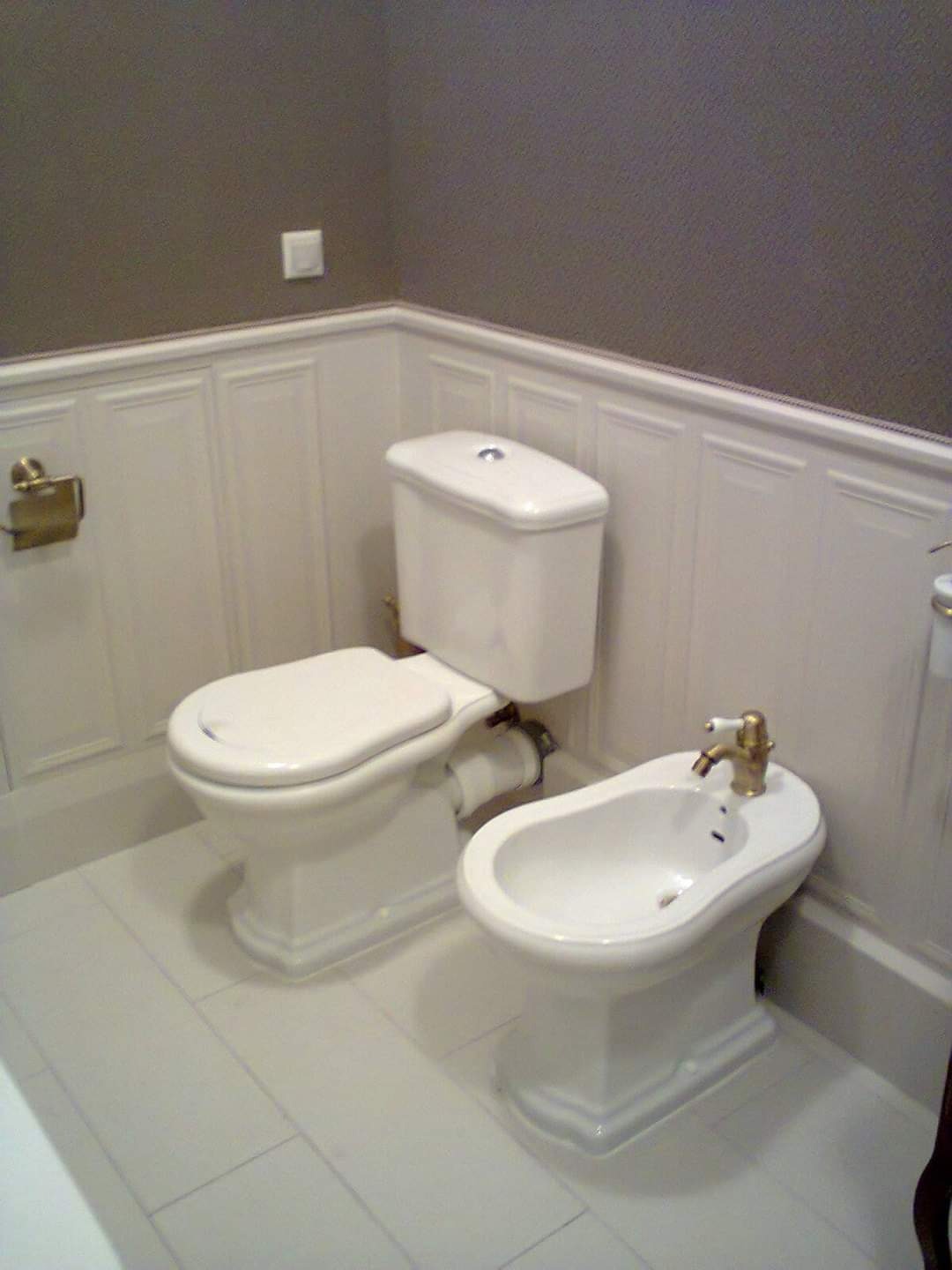 Biała toaleta i bidet w klasycznej łazience z panelami ściennymi i szarą tapetą. Złota armatura i akcesoria dodają elegancji. Widok z przodu, oświetlenie naturalne.