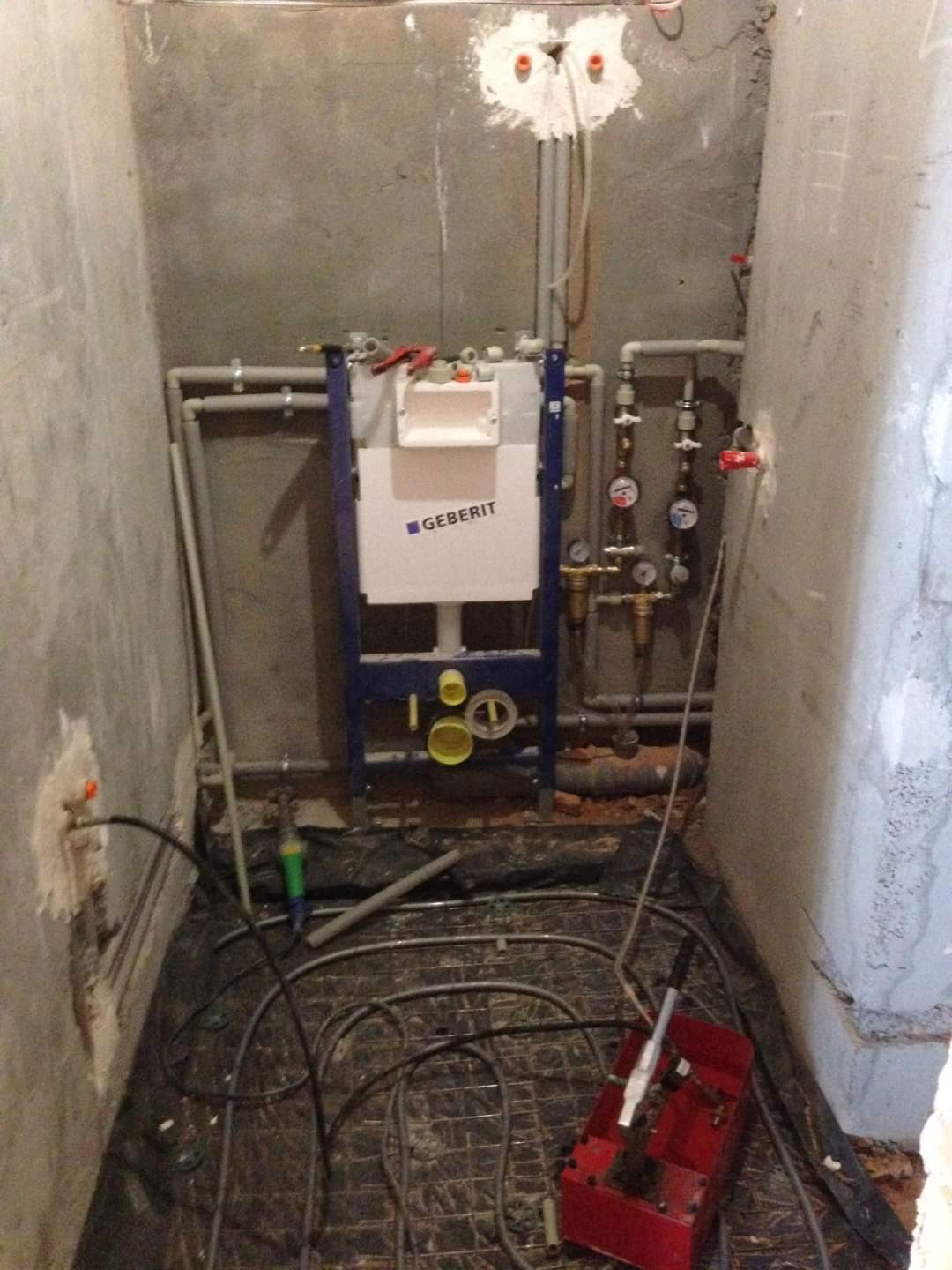 Montaż stelaża podtynkowego Geberit w surowej łazience z widoczną instalacją ogrzewania podłogowego i rurami doprowadzającymi wodę. Widoczne narzędzia hydrauliczne.