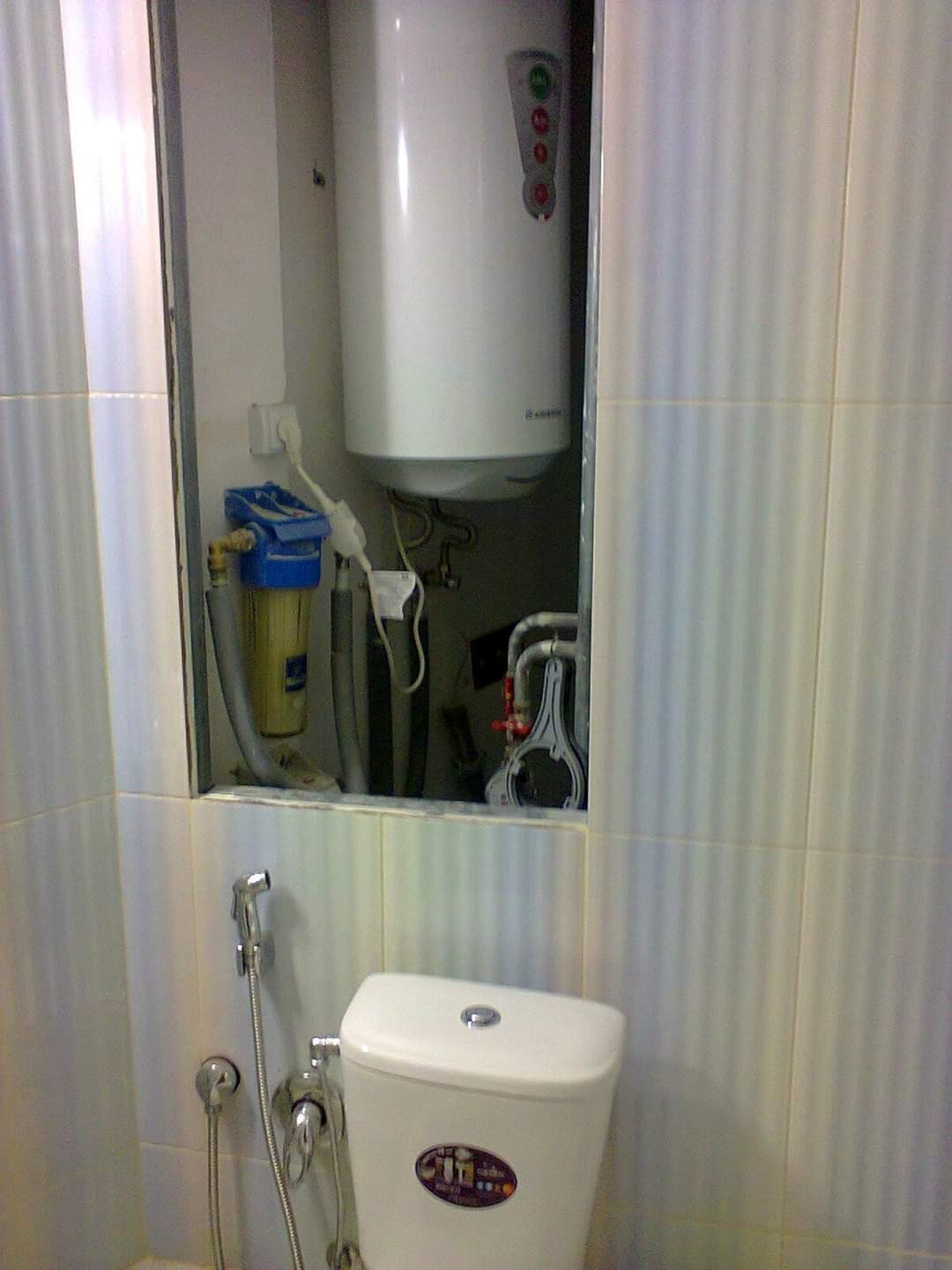 Biały, wiszący bojler Ariston w zabudowie, filtr wody i toaleta z bidetką w łazience z jasnymi, pionowymi płytkami. Widoczne elementy instalacji wodnej.