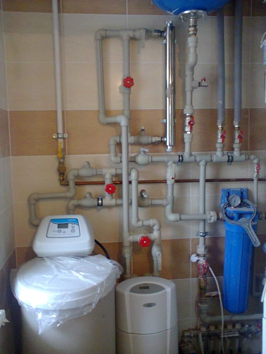 Złożona instalacja wodna z filtrami i zaworami w pomieszczeniu wyłożonym kafelkami. Widoczne rury, filtry wody (niebieski), zawory z czerwonymi pokrętłami i inne elementy instalacji.
