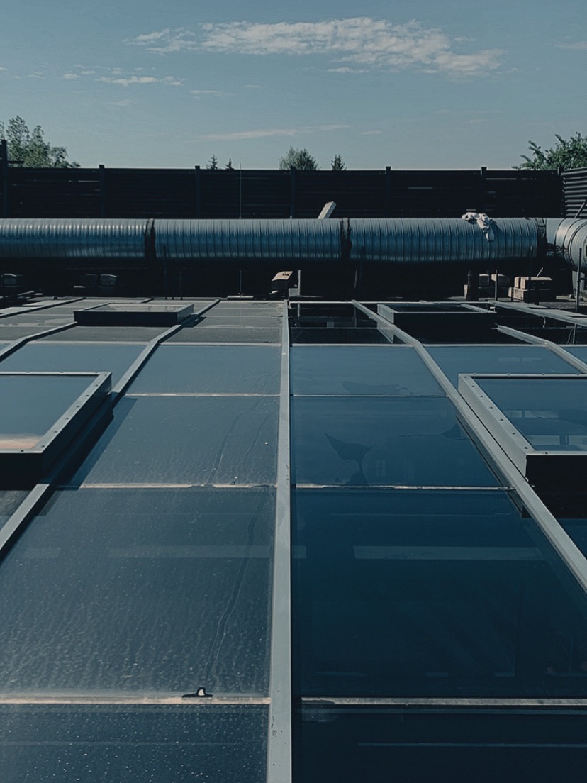 Szklany dach z metalową konstrukcją, widoczne zabrudzenia i zacieki. W tle instalacje wentylacyjne i fragment nieba z lekkimi chmurami. Niska perspektywa.