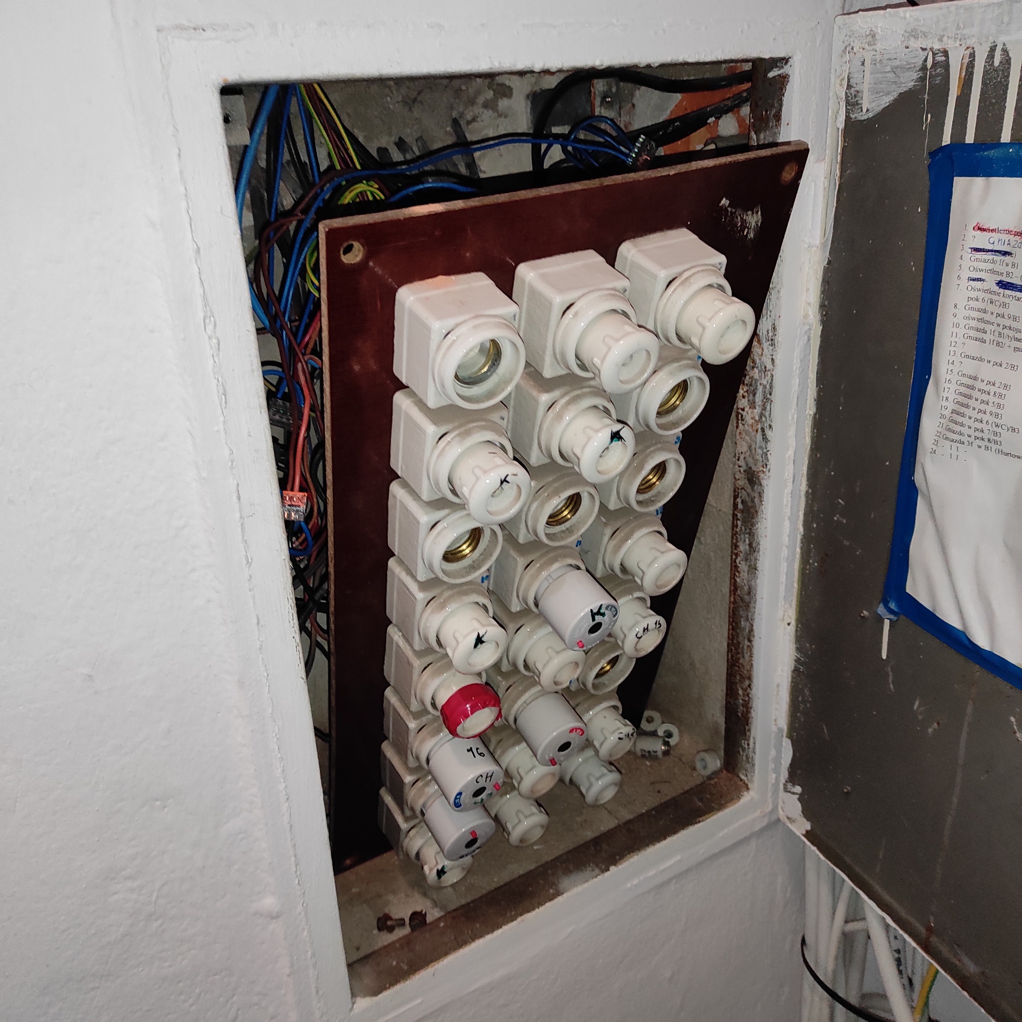 Otwarta skrzynka z bezpiecznikami topikowymi na brązowej płycie, widoczne kable i rozpiska obwodów. Jeden bezpiecznik z czerwoną nakładką. Wnętrze skrzynki z betonu.