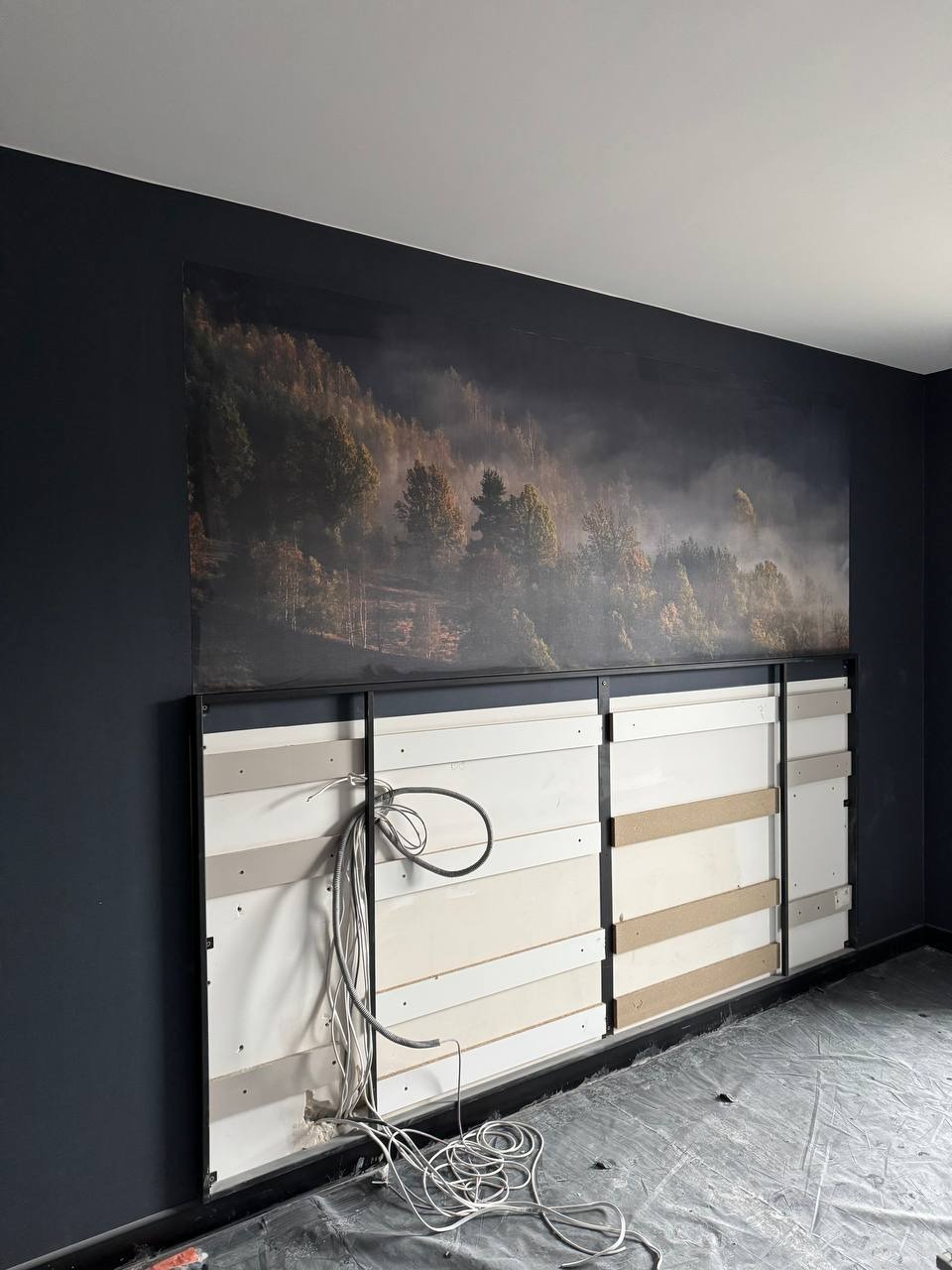 Montaż fototapety z motywem leśnym na stelażu z płyt, widoczne kable instalacji elektrycznej. Prace wykończeniowe w trakcie realizacji.